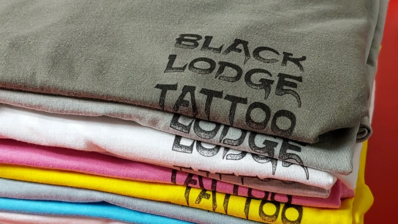 A stack of t-shirts in several colours printed with the Black Lodge Tattoo logo